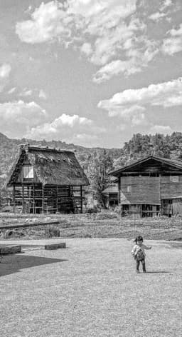 Photograph of a Young Child in the Foreground of Thatched Houses Beneath Clouds